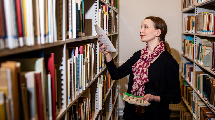 En kvinna står i ett bibliotek och håller i en bok.